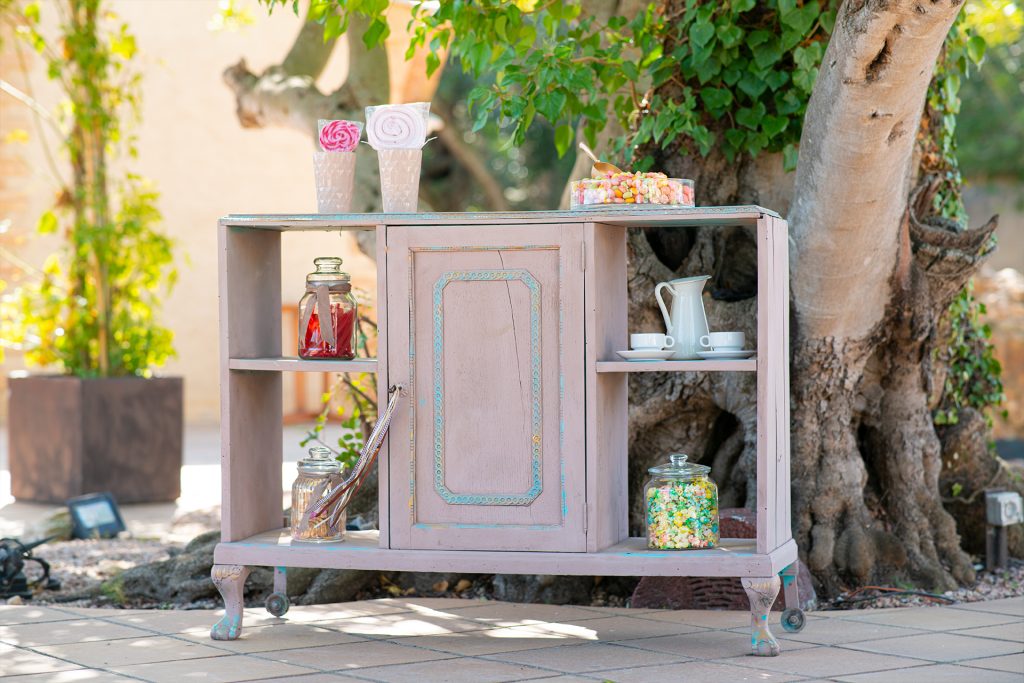 Pink chalk-painted cabinet set up outdoors as a sweets table with jars of sweets, a jug and cups